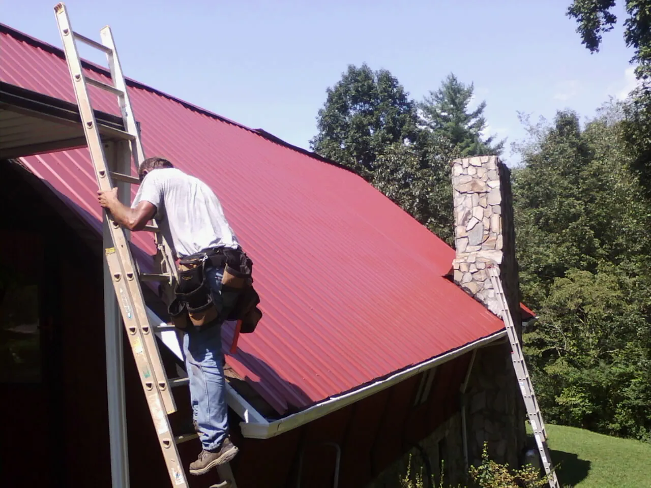 Roofer on a ladder beside a red metal roof and stone chimney — Soffit & Fascia Repair by Roofing Brothers in Hanoverton